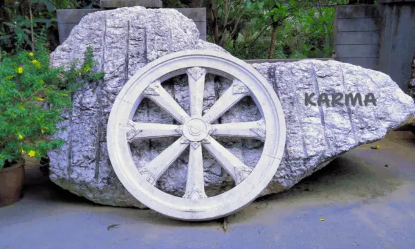 Photographie d'une roue ancienne en bois , posée sur un rocher gravé du mot Karma en pleine nature. Ce symbole illustre la loi de cause à effet et le cycle karmique.